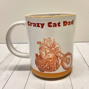 Peut inclure: Une tasse en céramique blanche avec une illustration brune d'un chat conduisant une moto. La tasse porte le texte "Crazy Cat Dad" en lettres orange.