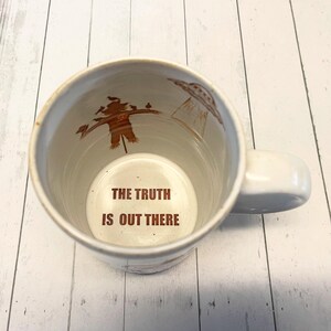 May include: White ceramic mug with a brown interior. The inside of the mug has a brown illustration of a scarecrow and a UFO. The bottom of the mug has the text "THE TRUTH IS OUT THERE" in brown.