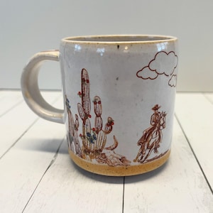 Może przedstawiać: Biały kubek ceramiczny z brązowym i białym wzorem przedstawiającym kowboja jadącego konno przez pustynny krajobraz z kaktusami. Kubek ma brązową obręcz i kropkowaną glazurę.
