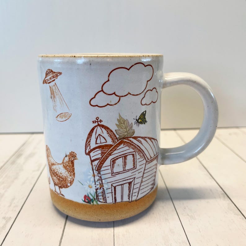 May include: White ceramic mug with a brown and orange design of a barn, clouds, a chicken, a butterfly, and a UFO. The mug has a brown rim and a brown base.
