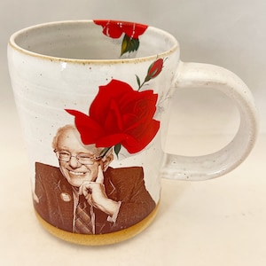May include: White ceramic mug with a large red rose and a portrait of a smiling man wearing glasses. The mug has a speckled finish and a tan base. The handle is curved and the interior is white.
