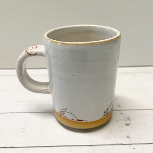 May include: White ceramic mug with a brown rim and a brown hand-painted design of a cat sleeping on a hill. The mug has a handle.