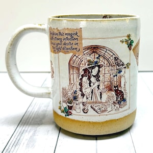 May include: A ceramic mug with a speckled off-white finish and a tan base. The illustration features a person in a hat, a black cat, a raven, and plants. Text reads: Perform this magick with strong intention. Focus your desire in the right direction.