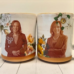 May include: Two white ceramic mugs with a brown rim and floral designs. Each mug features a portrait of a woman in a brown suit jacket. The mugs are on a white surface.