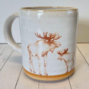May include: White ceramic mug with a brown moose design. The mug has a brown rim and a handle.