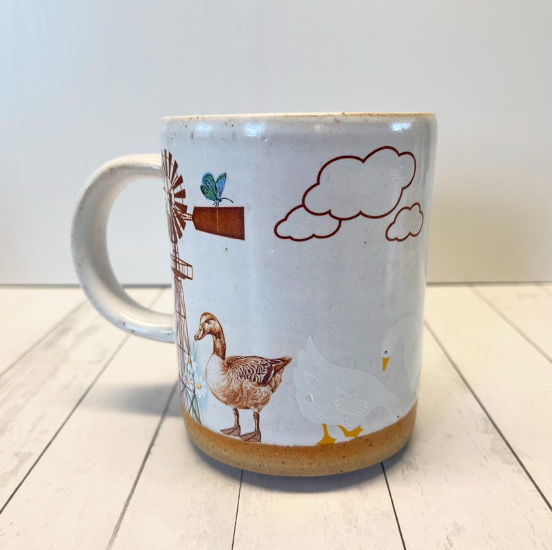 May include: White ceramic mug featuring a brown windmill, a butterfly, clouds, and two ducks. The duck on the left is brown and the duck on the right is white.