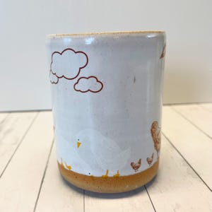 May include: White ceramic mug with a brown rim and a brown, yellow, and white design of clouds and chickens. The mug has a rustic, handmade look.