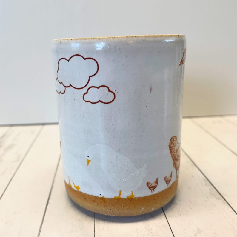 May include: White ceramic mug with a brown rim and a brown, yellow, and white design of clouds and chickens. The mug has a rustic, handmade look.