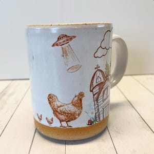 May include: White ceramic mug with a brown illustration of a farmhouse, chickens, and a flying saucer. The mug has a brown rim and a white handle.