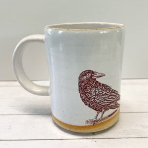 May include: White ceramic mug with a brown crow illustration. The mug has a rounded shape and a wide handle.