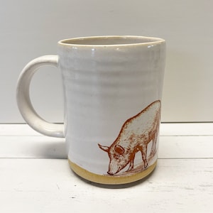 Könnte beinhalten: Weiße Keramiktasse mit einer braunen Illustration eines Schweins auf der Seite. Die Tasse hat einen gelben Rand und einen Henkel.