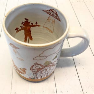 May include: White ceramic mug with a brown handle. The mug features a brown and white illustration of a scarecrow, birds, a barn, clouds, and a UFO.