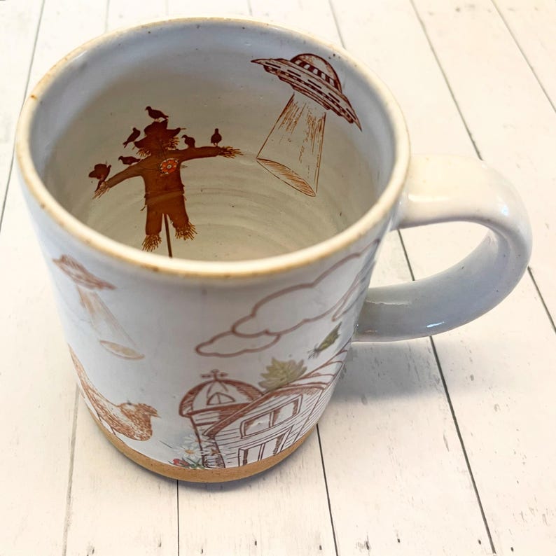 May include: White ceramic mug with a brown handle. The mug features a brown and white illustration of a scarecrow, birds, a barn, clouds, and a UFO.