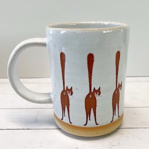 May include: A ceramic mug with a white speckled glaze and a tan base. The mug features three brown cat illustrations with their tails raised. The handle is white and rounded. A fun and quirky addition to any kitchen.