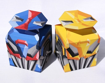Può includere: Due scatole di origami, una blu e una gialla, con disegni geometrici che ricordano i volti dei robot. Le scatole sono aperte e hanno un effetto 3D.