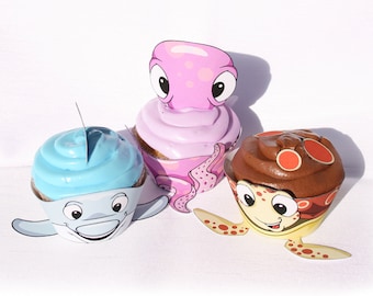 May include: Three decorated cupcakes with frosting. The cupcake on the left has blue frosting and a dolphin design. The cupcake in the middle has purple frosting and an octopus design. The cupcake on the right has brown frosting and a turtle design.