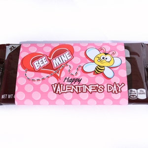 May include: A pink and white Valentine's Day chocolate bar wrapper with a red heart that says "Bee Mine" and a cartoon bee. The wrapper also says "Happy Valentine's Day".