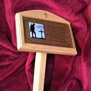 Op de afbeelding: Een houten gedenkplaat met een bronzen inzetstuk met een silhouet van een soldaat die voor een kruis staat. De plaat heeft een citaat over het gedenken van de overledenen: „Zij zullen niet oud worden, zoals wij die achterblijven oud worden. De leeftijd zal hen niet vermoeien, noch zullen de jaren hen veroordelen. Bij de ondergang van de zon en in de ochtend zullen wij hun gedenken.“