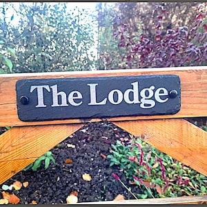 Könnte beinhalten: Ein Holztor mit einem schwarzen Schieferschild, auf dem "The Lodge" steht. Das Schild ist mit zwei silbernen Schrauben am Tor befestigt. Das Tor ist aus Holz und hat eine braune Farbe. Das Tor ist offen und im Hintergrund befindet sich ein grüner Busch.