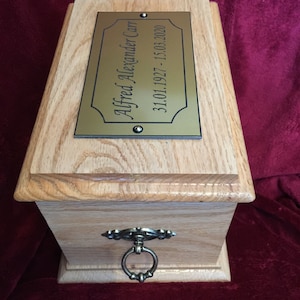 Op de afbeelding: Een houten crematie urn met een messingplaat met de tekst "Alfred Alexander Carr 31.01.1927 - 15.03.2020".