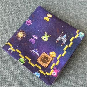 May include: A dark blue fabric square with a yellow border and a pixelated space theme. The fabric features images of spaceships, aliens, and stars. A gold label with the text "L.A.S. The Land of Art & Style" is in the center of the fabric.