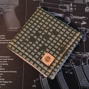 Peut inclure: Une pochette en tissu vert et beige avec une bordure cousue et un petit patch en cuir brun avec le texte "The L.A.S. & Sons" en lettres dorées.