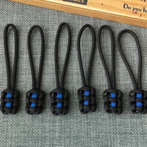 Op de afbeelding: Zes zwarte ritsen met blauwe strepen. De ritsen zijn gemaakt van paracord en hebben een knoop aan de onderkant.