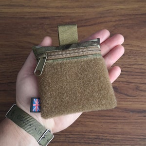 Könnte beinhalten: Eine kleine, braune Reißverschlusstasche mit Tarnmuster und Klettverschluss. Die Tasche ist aus weichem, strukturiertem Stoff gefertigt und hat eine Schlaufe zum Befestigen an einem Gürtel oder Rucksack.