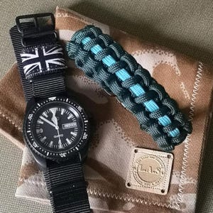 May include: A black wristwatch with a black nylon strap and a Union Jack patterned strap. The watch has a black face with white markings and a date window. The watch is on a brown camouflage fabric surface with a black and blue paracord bracelet and a leather patch with the text "The Tailor's  Ego & Co."