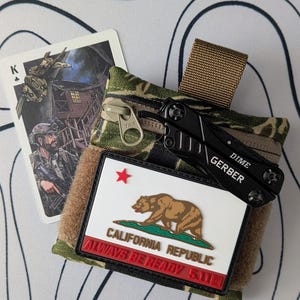 Puede incluir: Una funda con estampado de camuflaje con una multiherramienta Gerber Dime negra y una carta de juego con una ilustración militar. La funda tiene un parche blanco con el emblema de la República de California y el texto "ALWAYS BE READY 5.11®".