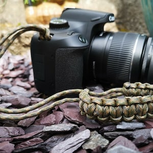 Könnte beinhalten: Eine schwarze DSLR-Kamera mit einem grünen und braunen Tarnmuster-Paracord-Handgelenkband. Die Kamera hat ein Objektiv angebracht und liegt auf einem Bett aus kleinen grauen Steinen.