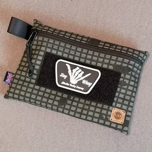 May include: An olive green pouch with a checkered pattern, black zip, and a patch reading "Stay Wavy". The pouch features a Union Jack tag and a small leather tag with "L.A.S.". The design is a practical accessory, suitable for various uses.