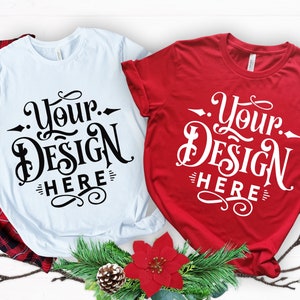 Könnte beinhalten: Zwei T-Shirts, eines weiß und eines rot, mit dem Text "Your Design Here" in einer dekorativen Schriftart. Die Shirts werden mit Weihnachtsdekorationen präsentiert, darunter ein rot kariertes Hemd, Tannenzweige und ein Weihnachtsstern.