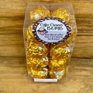 May include: A clear plastic container filled with several gold-wrapped coffee creamer bombs. The label reads "Coffee Creamer BOMB" with instructions to pour 8 oz of coffee over 1 or 2 bombs. The bottom label says "French Vanilla."