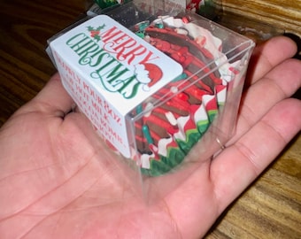 Może przedstawiać: Przezroczyste plastikowe pudełka zawierające bomby kakaowe o tematyce świątecznej. Każde pudełko ma etykietę z napisem "Merry Christmas" z ilustracją czapki Świętego Mikołaja. Bomby kakaowe są czerwone, białe i zielone, z świątecznym wzorem.