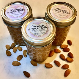 May include: Three glass jars of homemade almond butter with silver lids. The jars have a diamond pattern and labels that read "Homemade Almond Butter, Extra Creamy." Almonds are scattered around the jars. One jar is strawberry flavored.