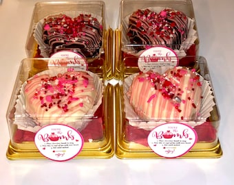 May include: Four heart-shaped chocolate bombs in clear plastic containers. Two are dark chocolate with pink and red sprinkles, and two are white chocolate with pink drizzle and sprinkles. Each container has a sticker that says "You're the Bomb".
