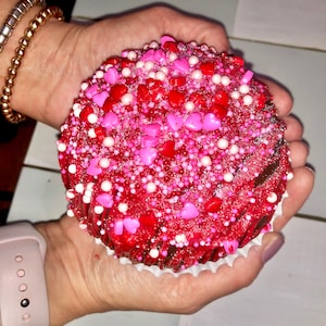 May include: A large, red cupcake with a generous topping of sprinkles and heart-shaped candies. The cupcake is held in cupped hands, showcasing the festive decorations. The cupcake sits in a white paper liner.