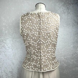 Pode incluir: Um top sem mangas com fundo bege e detalhes de contas brancas. O top tem um fecho de correr nas costas.
