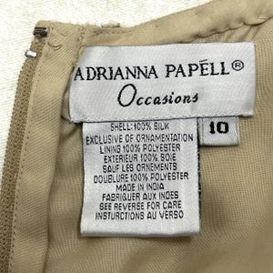 Pode incluir: Uma etiqueta branca com texto preto detalhando o conte&uacute;do do tecido e as instru&ccedil;&otilde;es de cuidado para um vestido. A etiqueta diz "Adrianna Papell Occasions", "Shell: 100% Silk", "Exclusive of ornamentation", "Lining 100% Polyester", "Exterieur 100% Soie", "Sauf les ornements", "Doublure 100% Polyester", "Made in India", "Fabriquer aux Indes", "See reverse for care instructions au verso". A etiqueta tamb&eacute;m tem uma etiqueta branca com o n&uacute;mero "10" impresso em preto.