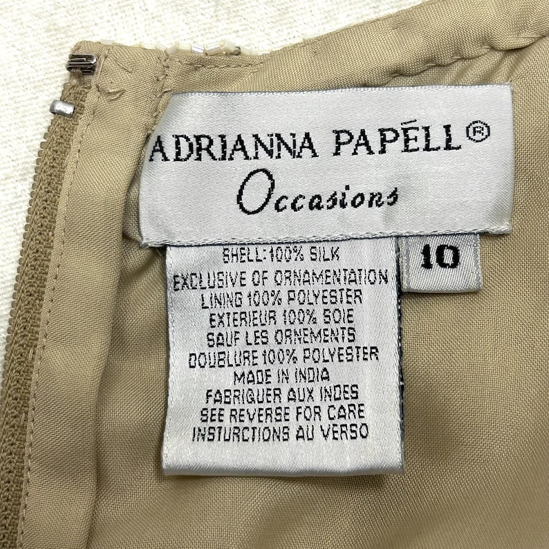 Pode incluir: Uma etiqueta branca com texto preto detalhando o conte&uacute;do do tecido e as instru&ccedil;&otilde;es de cuidado para um vestido. A etiqueta diz "Adrianna Papell Occasions", "Shell: 100% Silk", "Exclusive of ornamentation", "Lining 100% Polyester", "Exterieur 100% Soie", "Sauf les ornements", "Doublure 100% Polyester", "Made in India", "Fabriquer aux Indes", "See reverse for care instructions au verso". A etiqueta tamb&eacute;m tem uma etiqueta branca com o n&uacute;mero "10" impresso em preto.