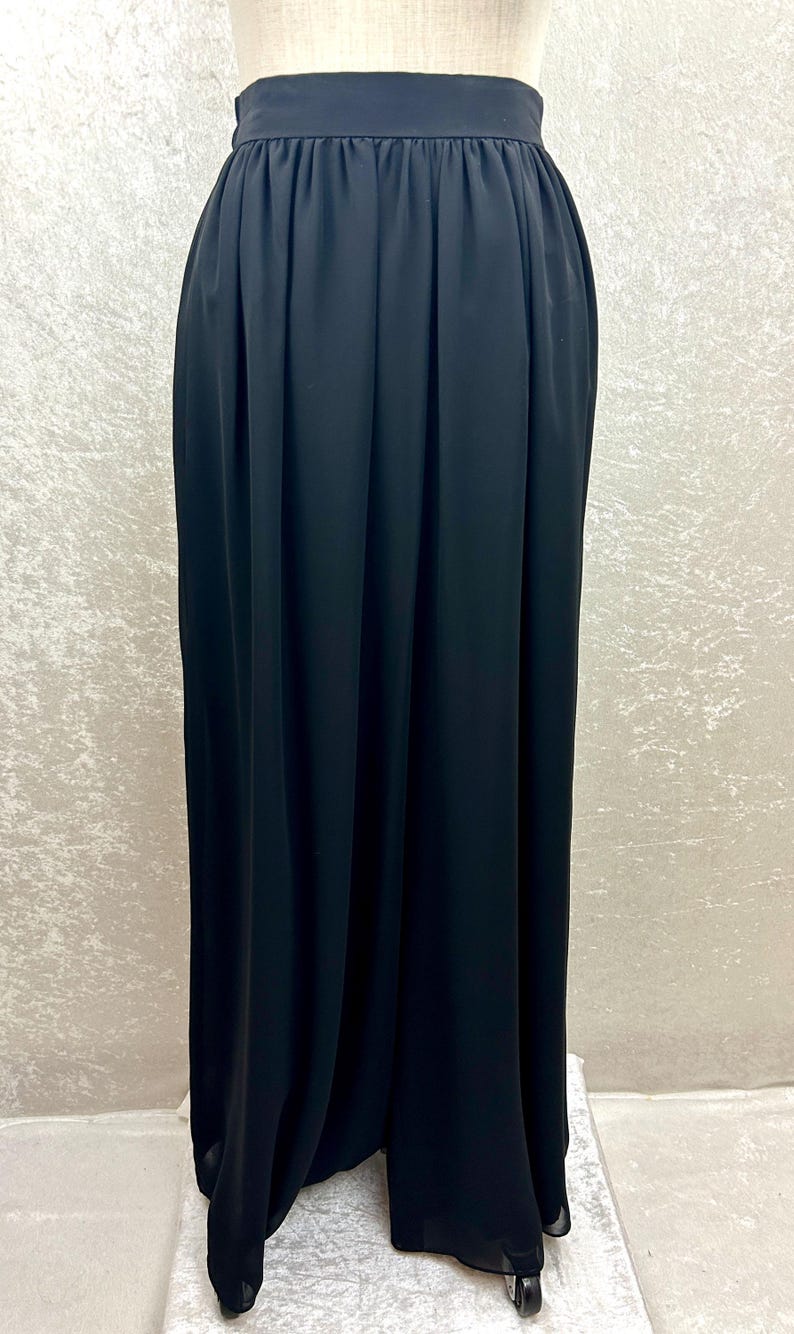 Puede incluir: Una falda larga negra con cintura alta y tejido fluido. La falda se muestra en un maniqu&iacute;, destacando su dise&ntilde;o plisado y su elegante ca&iacute;da. El color oscuro y el dise&ntilde;o sencillo sugieren versatilidad.