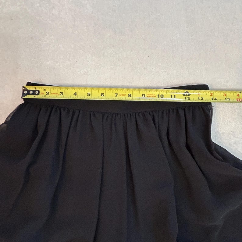 Puede incluir: Falda negra con cintura fruncida, medida con una cinta m&eacute;trica amarilla. La cintura de la falda mide aproximadamente 33 cm de ancho. La tela parece ser un material ligero y fluido.