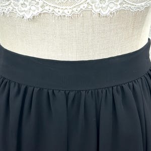 Puede incluir: Falda negra con una cintura ancha y un dise&ntilde;o plisado y fluido. La falda est&aacute; hecha de una tela ligera y se muestra en un maniqu&iacute;. La parte superior del maniqu&iacute; es visible, con un detalle de encaje blanco.