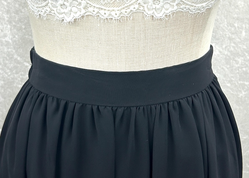 Puede incluir: Falda negra con una cintura ancha y un dise&ntilde;o plisado y fluido. La falda est&aacute; hecha de una tela ligera y se muestra en un maniqu&iacute;. La parte superior del maniqu&iacute; es visible, con un detalle de encaje blanco.