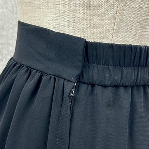 Puede incluir: Primer plano de una falda negra con cintura el&aacute;stica y cierre de cremallera. La tela de la falda parece ligera y fluida, con sutiles detalles de pliegues. La prenda se muestra sobre un maniqu&iacute;.