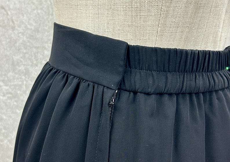 Puede incluir: Primer plano de una falda negra con cintura el&aacute;stica y cierre de cremallera. La tela de la falda parece ligera y fluida, con sutiles detalles de pliegues. La prenda se muestra sobre un maniqu&iacute;.