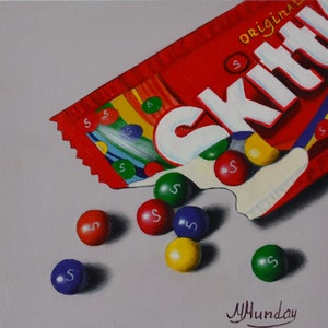Op de afbeelding: Een rode en witte zak Skittles snoepjes met het woord "Skittles" erop gedrukt. De zak is open en er vallen een aantal Skittles snoepjes uit. De snoepjes zijn rood, oranje, geel, groen en paars.