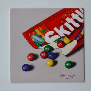 Op de afbeelding: Een schilderij van een gemorst zakje Skittles snoep. Het zakje is rood en wit met het woord "Skittles" erop gedrukt. Het snoep ligt verspreid over een lichtgrijze ondergrond. Het snoep is rood, oranje, geel, groen en paars.