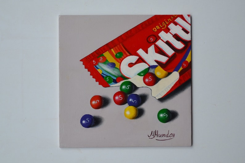 Op de afbeelding: Een schilderij van een gemorst zakje Skittles snoep. Het zakje is rood en wit met het woord "Skittles" erop gedrukt. Het snoep ligt verspreid over een lichtgrijze ondergrond. Het snoep is rood, oranje, geel, groen en paars.